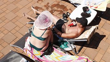 Nainen istuu aurinkohattu päässään ja hänen vieressään aurinkotuolilla makoileva mies hörppii limua pillillä.